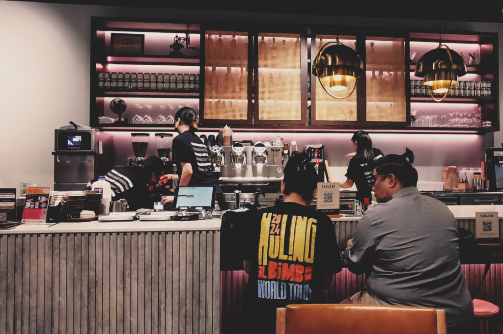 Busy coffee shop scene with baristas working espresso machines, warm lighting, and customers enjoying drinks at counter.