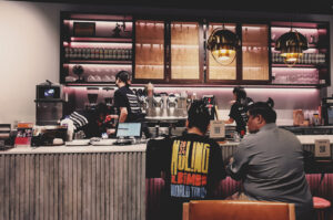 Busy coffee shop scene with baristas working espresso machines, warm lighting, and customers enjoying drinks at counter.