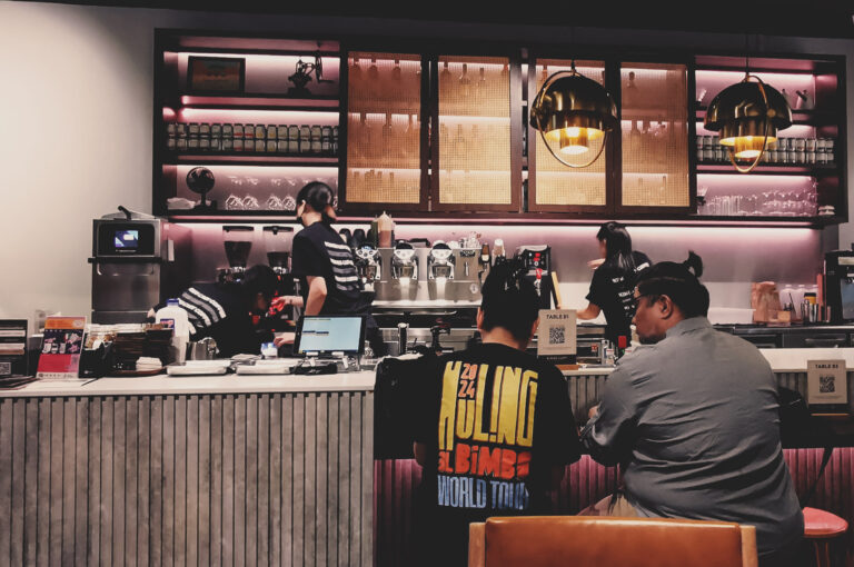 Busy coffee shop scene with baristas working espresso machines, warm lighting, and customers enjoying drinks at counter.