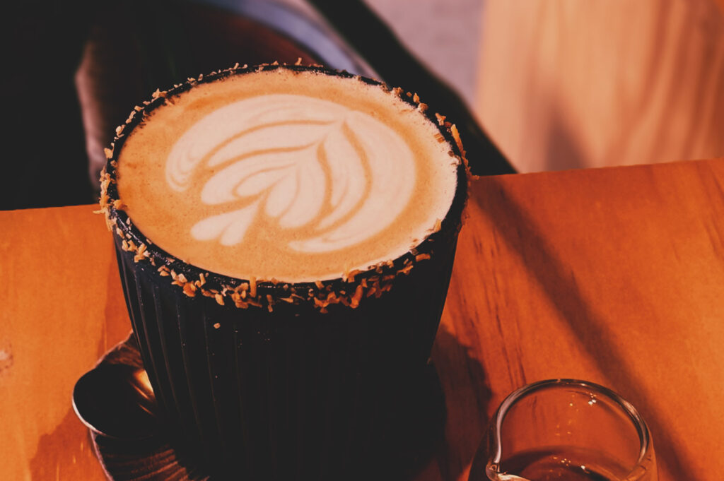 Latte in black ceramic cup with leaf latte art and toasted coconut rim, served on coaster with spoon.