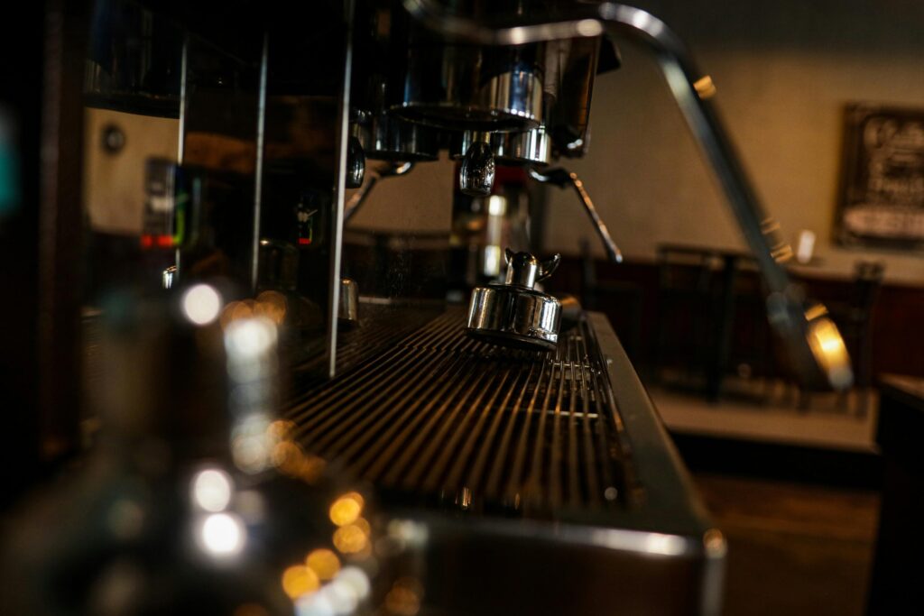 Cafe Photographer – 4 A shallow depth of field focuses sharply on the metallic drip tray of an espresso machine, where a tamper rests against the gleaming stainless steel lines. Foreground bokeh adds a shimmering golden texture, contrasting with the blurred, warmly lit cafe interior visible in the background.