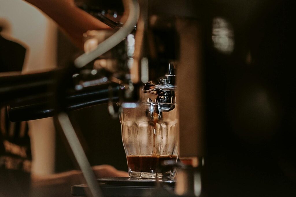 A steady stream of dark espresso drips from a chrome spout into a faceted glass cup. The shallow depth of field blurs the foreground machinery, drawing the eye directly to the rich coffee extraction at the center.