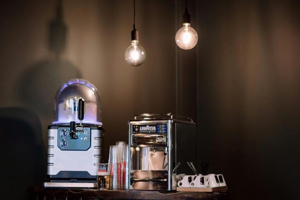 Two hanging Edison bulbs cast a warm glow over a counter featuring a Lavazza Espresso Point machine and a beverage dispenser with a domed top. The lighting creates moody shadows on the dark wall behind, highlighting the metallic surfaces and the neat arrangement of cups and stirrers.