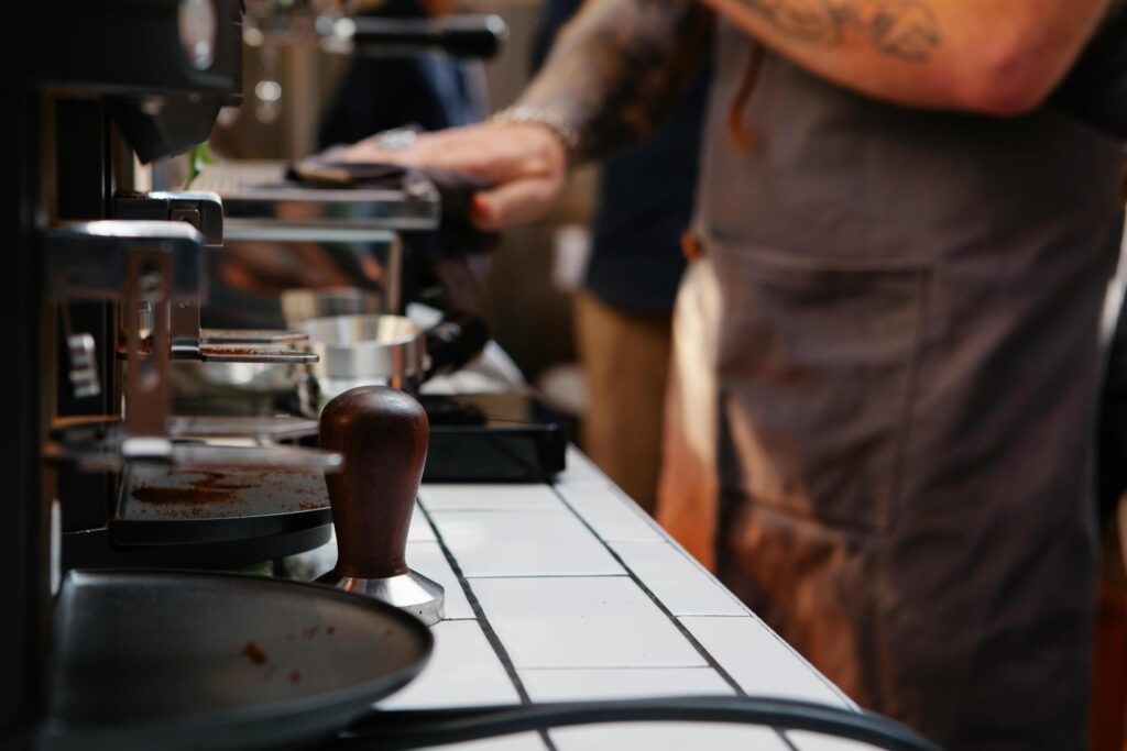 A wooden-handled tamper sits on a white tiled counter alongside scattered coffee grounds, anchored sharply in the foreground against the machinery. Behind it, the blurred figure of a tattooed barista in a gray apron tends to the workspace, creating a candid "behind-the-scenes" atmosphere.