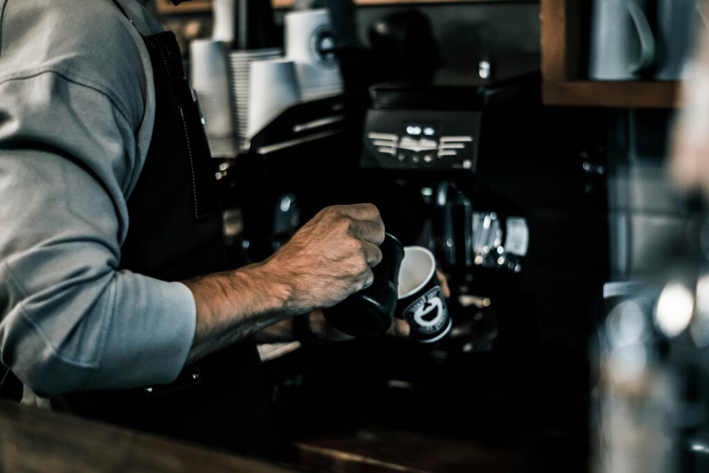 A barista clad in a grey shirt and dark apron stands poised with a black pitcher, preparing to pour into a branded paper cup held in their other hand. The composition focuses on the tactile details of the coffee-making process, with stacks of cups and espresso machinery blurring into the dark, moody background.