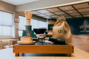 A wooden display tray neatly presents an assortment of artisanal coffee and tea essentials, including a metal portafilter, a bamboo matcha whisk, and a bowl of roasted beans. The set is highlighted by "Suzuki Gourmet Coffee" branding on a burlap sack and paper cup, placed against the blurred background of a modern cafe interior.
