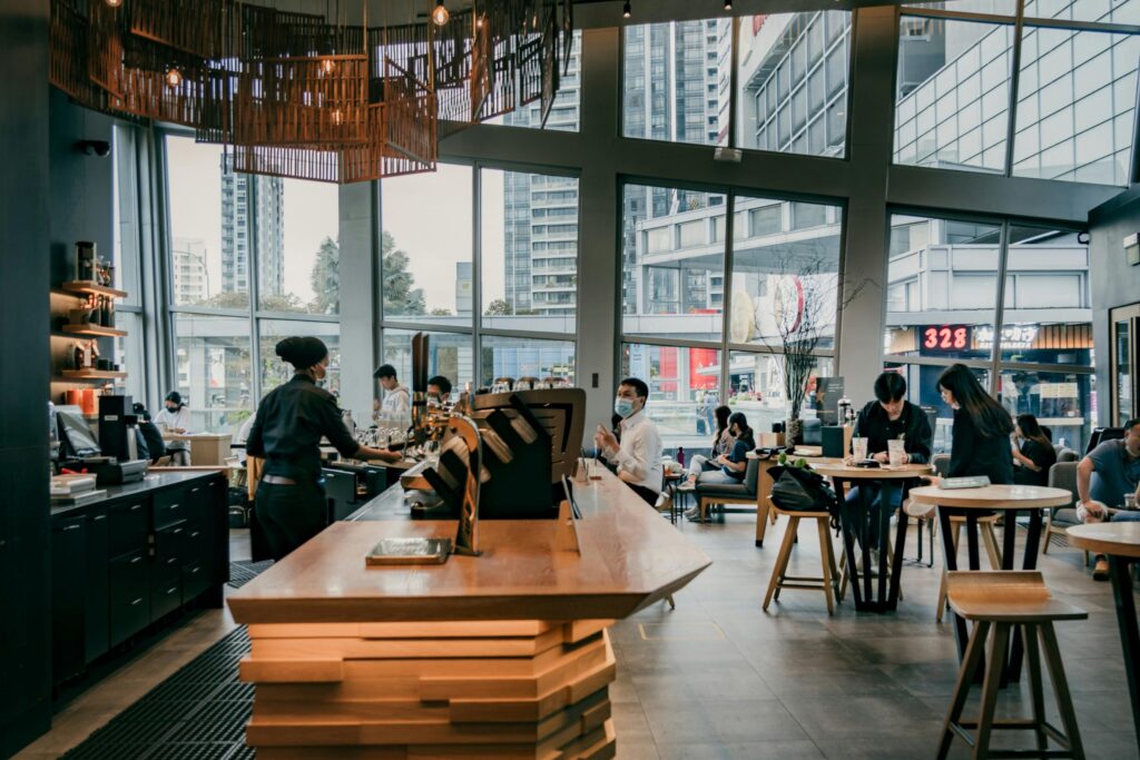 Expansive floor-to-ceiling windows wrap around this modern cafe, flooding the space with natural light and providing views of the surrounding city skyline. Underneath a large, intricate wooden chandelier, a barista works behind a prominent service counter while customers sit at scattered tables throughout the interior.