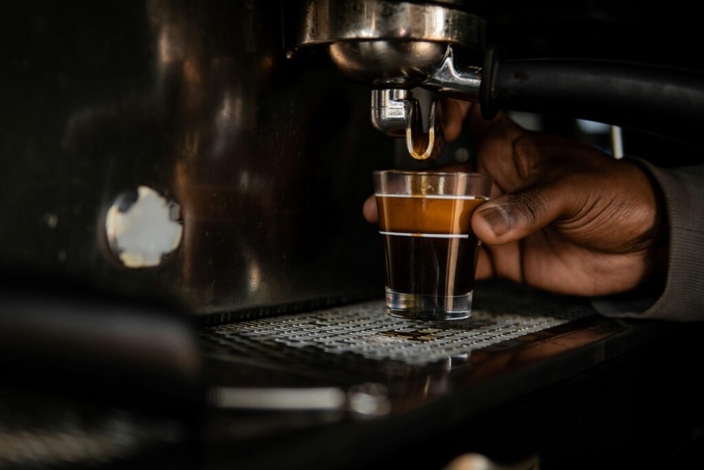 Slideshow – 1 A close-up captures a hand carefully holding a small glass measuring cup beneath the spout of a stainless steel espresso machine. Rich, dark coffee topped with a thick layer of golden crema streams steadily into the glass.