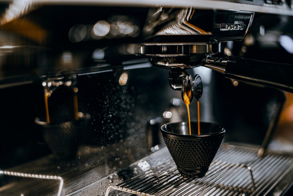 Slideshow – 4 Twin streams of golden-brown espresso pour from a double-spout portafilter into a black, textured cup sitting on a wire drip tray. The polished metallic backsplash acts as a mirror, clearly reflecting the extraction process and the soft bokeh lights from the dark background.