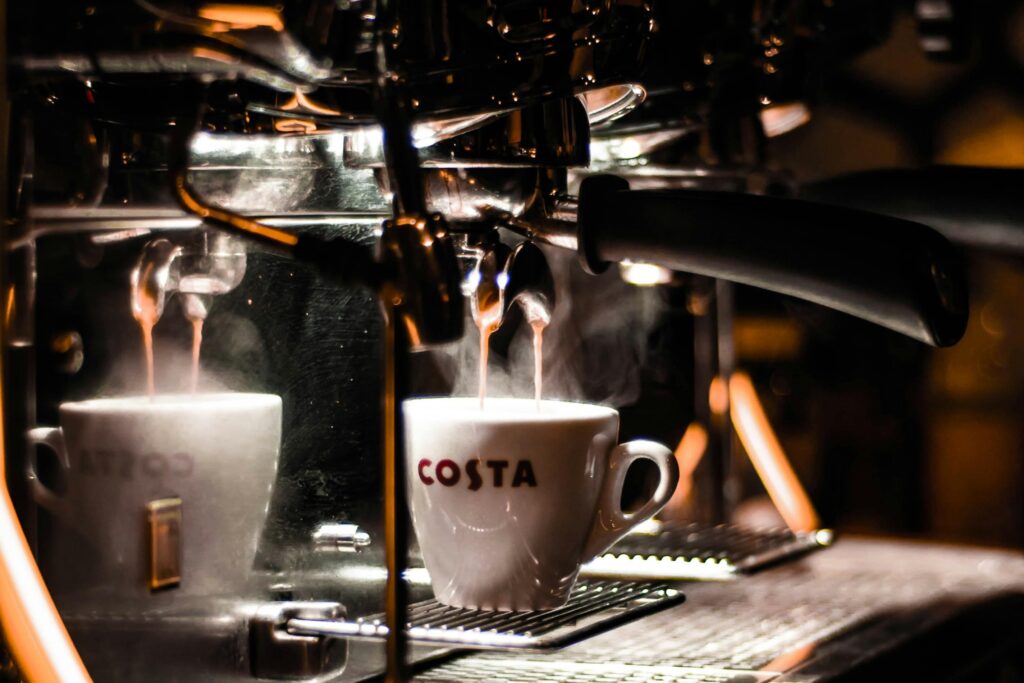 Slideshow – 5 Steaming espresso pours in dual streams from a chrome portafilter into a white ceramic cup branded with the red "COSTA" logo. Warm backlighting dramatizes the scene, catching the swirling vapor rising from the cup and creating deep reflections on the machine's polished metallic exterior.