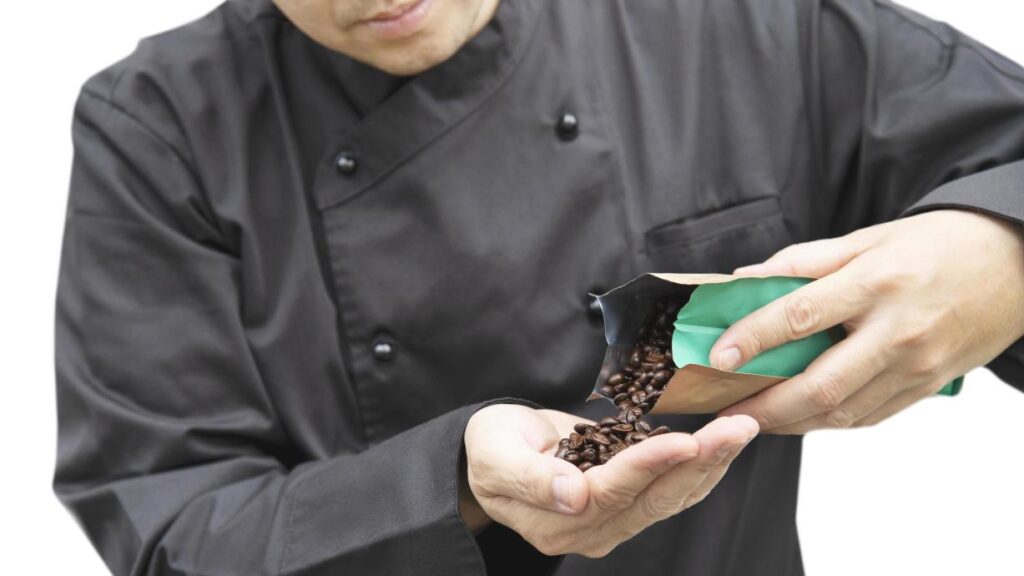 The Approach A person wearing a black chef's jacket pours dark roasted coffee beans from a package into their open hand. The close-up shot highlights the texture of the beans as they are apparently being inspected against a white background.