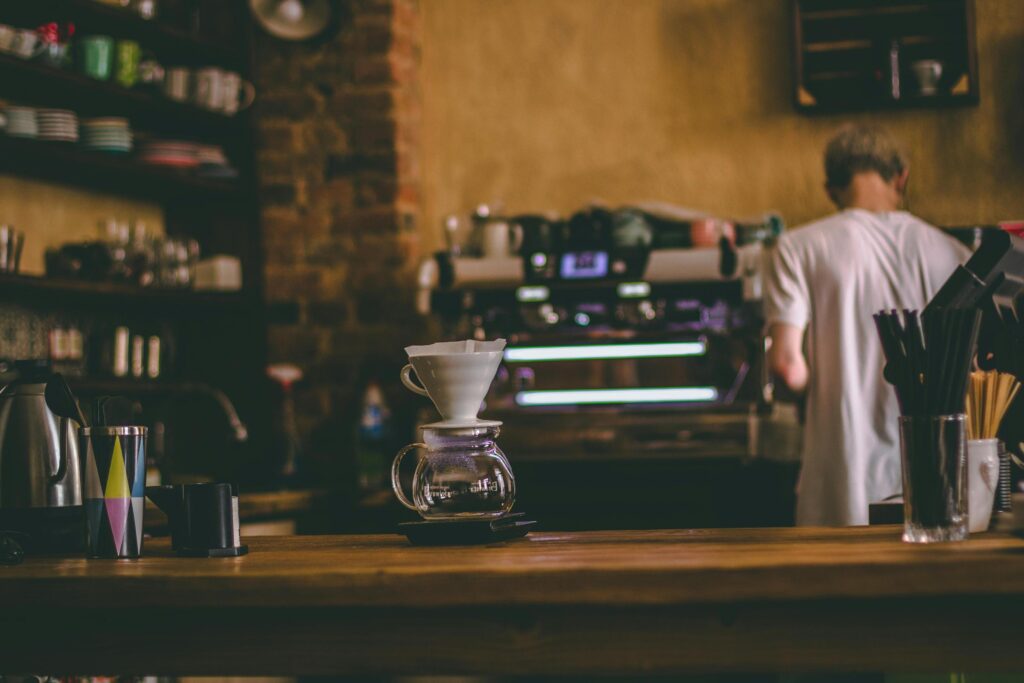 A glass pour-over coffee maker with a white ceramic dripper sits in sharp focus on a wooden counter, serving as the centerpiece of this cozy scene. In the softly blurred background, a barista in a white t-shirt tends to an espresso machine amidst brick walls and shelves of mugs.