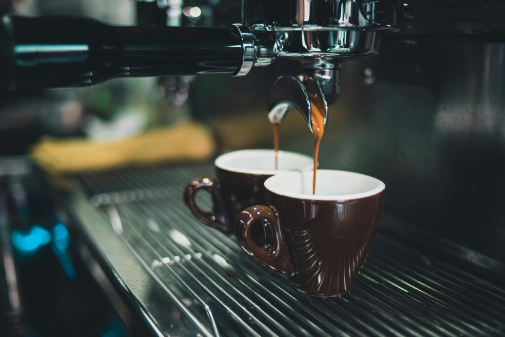 An espresso machine pours two steady streams of hot coffee into a pair of dark brown cups positioned on a metal grate. The close-up shot captures the rich liquid flowing from the chrome portafilter, set against a blurred background that emphasizes the brewing process.