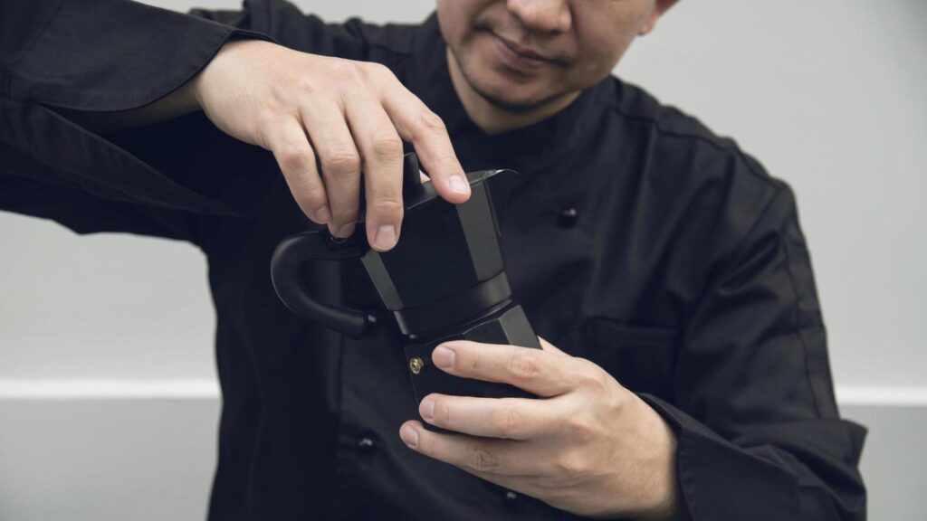 A man dressed in a black chef's jacket is shown holding a black Moka pot in front of a plain background. He uses both hands to carefully adjust or secure the top section of the espresso maker.
