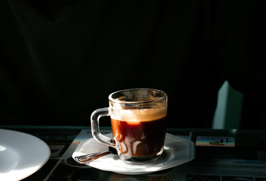 A small glass cup containing dark coffee with a layer of foam sits on a white saucer, accompanied by a silver spoon. Sharp, natural sunlight illuminates the beverage from the side, creating a high-contrast effect against the shadowed background.