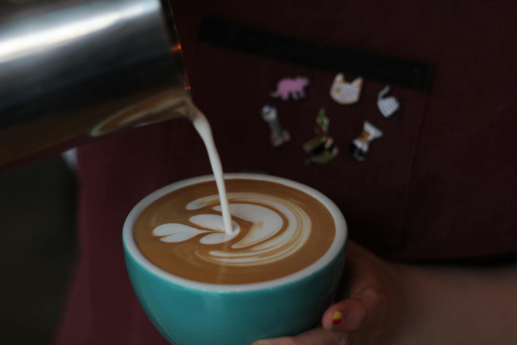 A barista carefully pours steamed milk from a silver pitcher into a teal cup, forming a white latte art pattern on the surface of the coffee. The person wears a maroon apron decorated with several small enamel pins, including cat and coffee-themed designs.