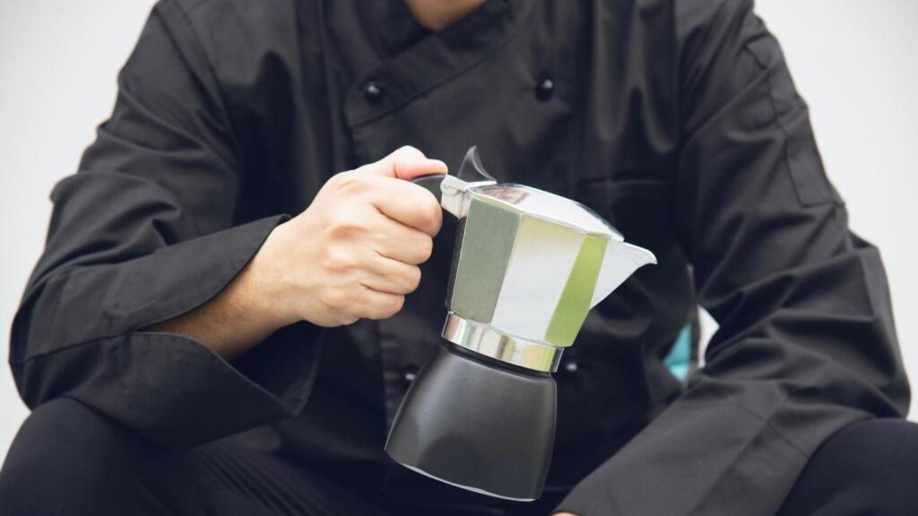 A person wearing a black chef's uniform holds a Moka pot that features a silver upper chamber and a black base. They are gripping the handle of the espresso maker, which is positioned centrally in the frame against a plain background.