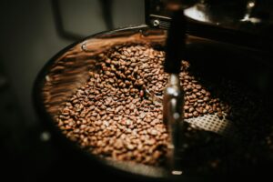 This close-up captures a large mound of roasted coffee beans swirling inside the metallic cooling tray of an industrial roaster. The scene is bathed in warm, dramatic lighting that accentuates the rich texture of the beans and the gleaming surfaces of the machinery.