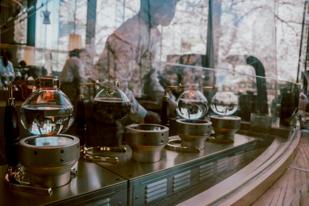 A row of elegant glass siphon coffee makers sits lined up on a metal counter, showcasing their spherical bowls and intricate brass fixtures. The scene is viewed through a reflective glass partition that overlays ghostly silhouettes of the outside trees and city streets onto the café interior.