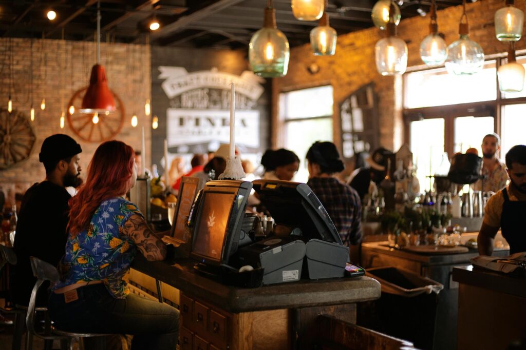 Warm hanging lights illuminate a bustling bar scene featuring exposed brick walls and rustic decor. In the foreground, patrons sit at the wooden counter near a service station, surrounded by a lively crowd enjoying the cozy atmosphere.