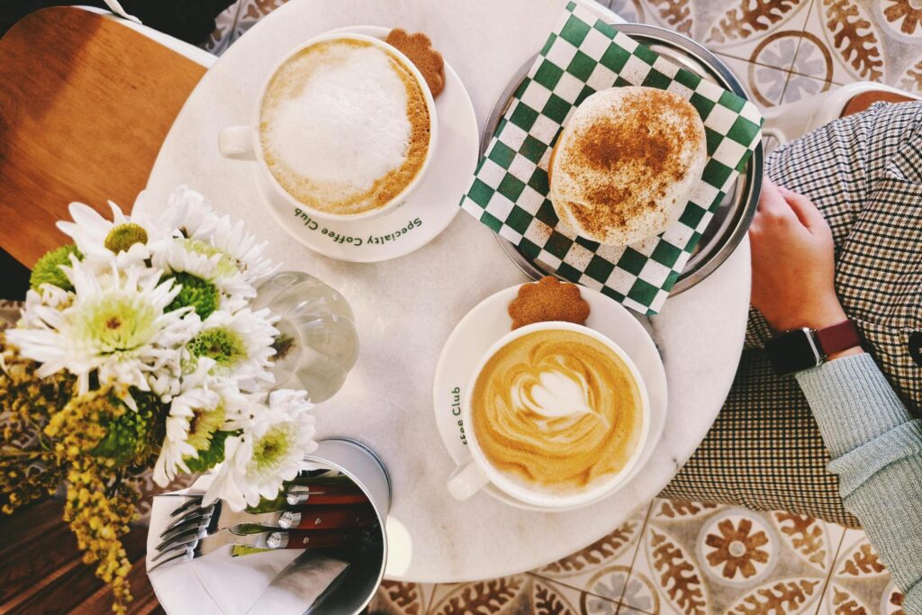 Captured from above, this cozy café scene features a round marble table holding two cups of coffee, a cinnamon-dusted pastry on a green checkered napkin, and a vase of white daisies. The aesthetic arrangement is framed by a patterned tile floor and a glimpse of a person in a plaid blazer enjoying the spread.