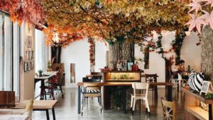 This cafe features a cozy, indoor autumn theme centered around a large artificial tree with vibrant orange and yellow leaves spreading across the ceiling. The space is filled with wooden tables, mismatched chairs, and warm lighting, creating an inviting atmosphere for visitors to relax or work.