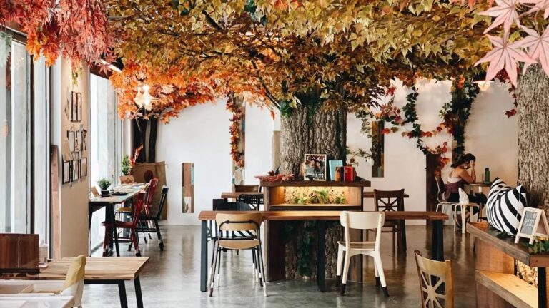 This cafe features a cozy, indoor autumn theme centered around a large artificial tree with vibrant orange and yellow leaves spreading across the ceiling. The space is filled with wooden tables, mismatched chairs, and warm lighting, creating an inviting atmosphere for visitors to relax or work.