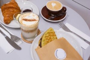 This image features a cozy cafe spread consisting of a croissant with butter and jam, a waffle sandwich, and two coffee drinks. The items are neatly arranged on a white table alongside cutlery and napkins, including a latte with heart-shaped foam and an iced coffee topped with cream.