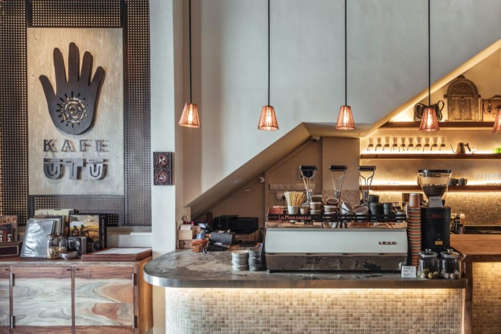 The image showcases the interior of Kafe Utu, featuring a rustic wooden sign with a hand logo and warm, low-hanging pendant lights. A sleek coffee bar equipped with a professional espresso machine sits beneath a sloped ceiling, complemented by earthy textures and mosaic tiling