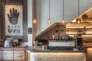 The image showcases the interior of Kafe Utu, featuring a rustic wooden sign with a hand logo and warm, low-hanging pendant lights. A sleek coffee bar equipped with a professional espresso machine sits beneath a sloped ceiling, complemented by earthy textures and mosaic tiling