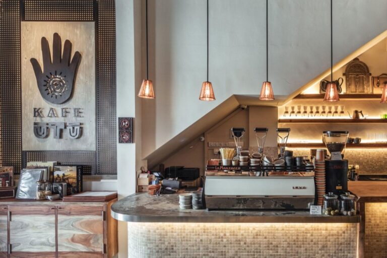 The image showcases the interior of Kafe Utu, featuring a rustic wooden sign with a hand logo and warm, low-hanging pendant lights. A sleek coffee bar equipped with a professional espresso machine sits beneath a sloped ceiling, complemented by earthy textures and mosaic tiling
