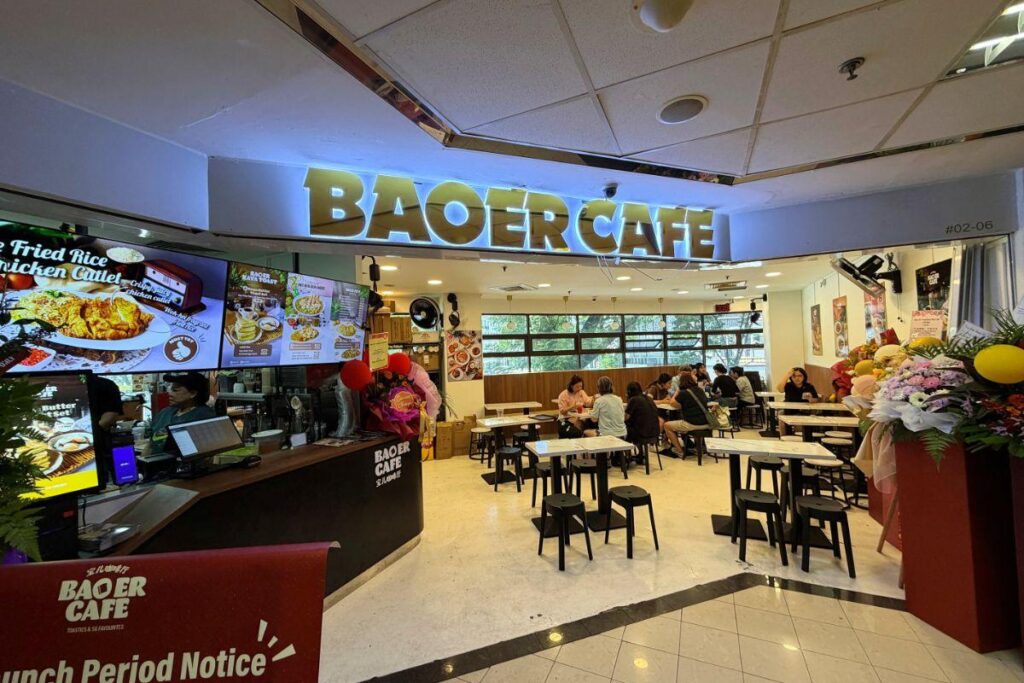 The image showcases the bright storefront of Baoer Cafe, featuring a large golden sign above an open seating area with customers. Digital menu boards display various food options next to the ordering counter, while white tables and black stools fill the indoor dining space.