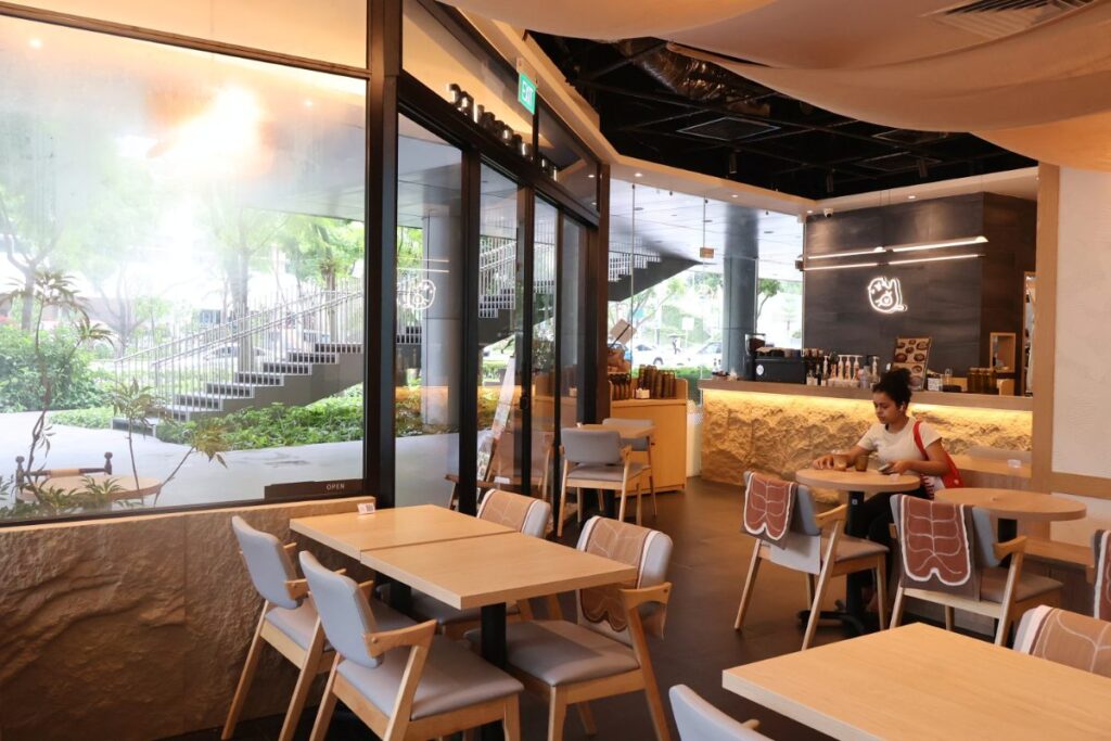 The interior of the cafe features light wood tables and grey chairs arranged near large glass windows that look out onto a green landscape. A service counter with warm under-lighting is visible in the background, where a customer sits at a nearby table in the minimalist, modern space.