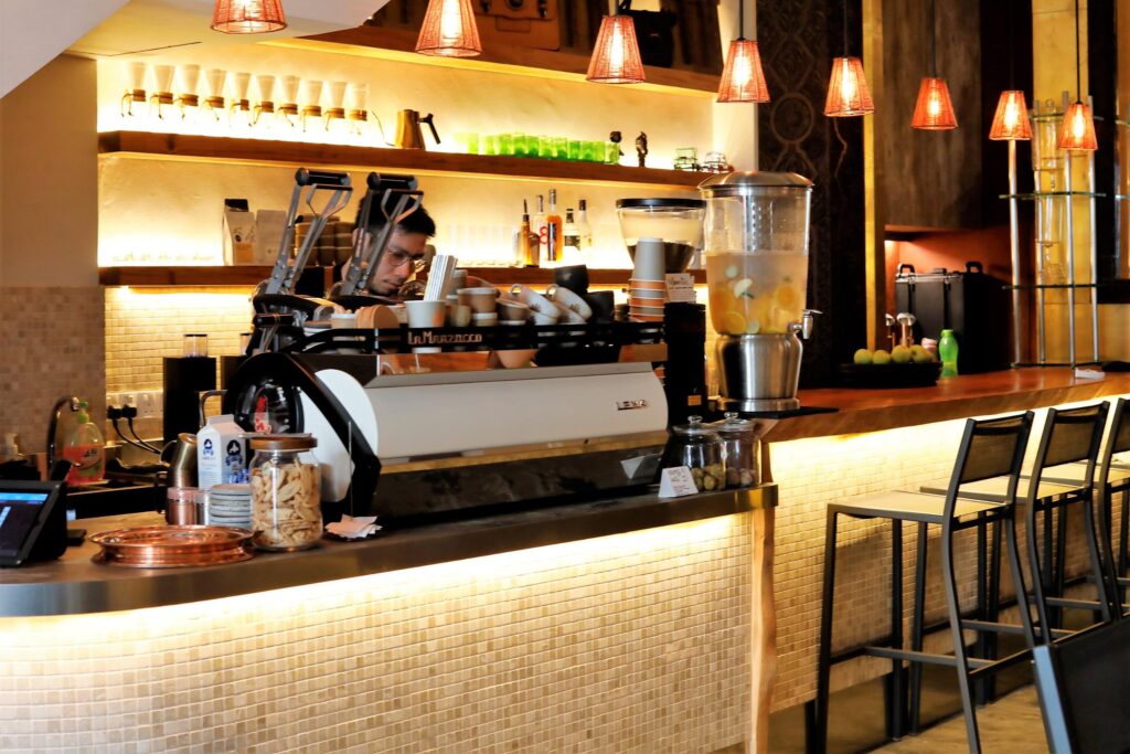 A barista works behind a modern coffee bar illuminated by warm pendant lights and an under-counter glow. The setting features a professional espresso machine, shelves stocked with glassware, and a row of tall bar stools along a tiled counter.