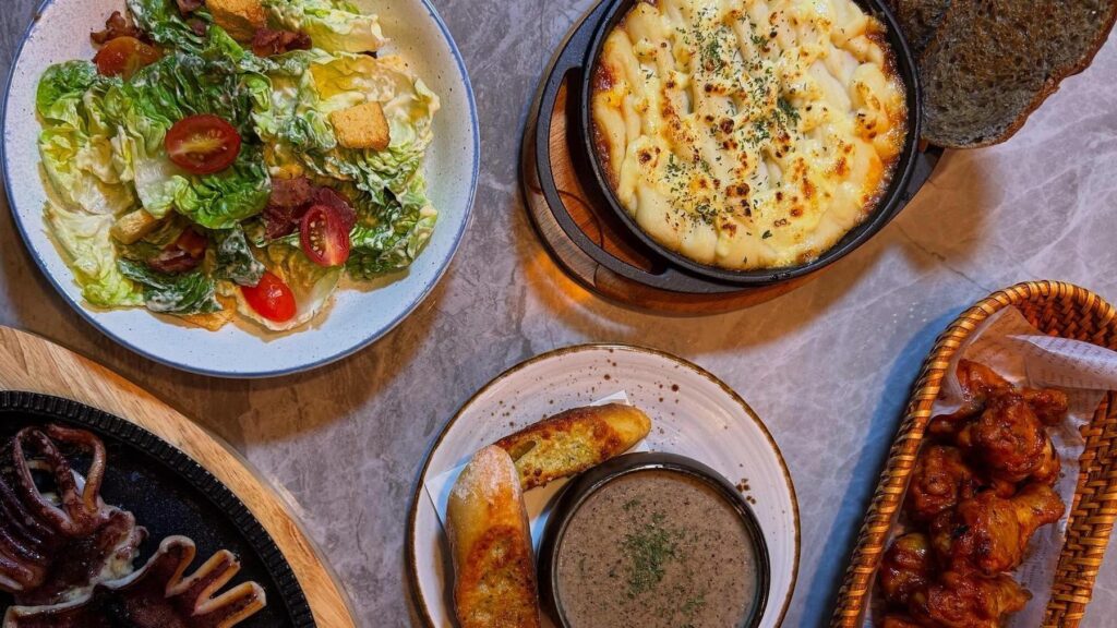This top-down shot displays a variety of appetizing dishes, including a fresh Caesar salad, a cheesy skillet pasta, and crispy fried chicken wings. The spread is rounded out with a creamy bowl of soup served with toasted bread and a grilled squid dish, all set against a grey marble-style background.