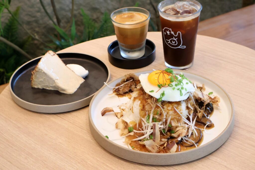 A spread of cafe items sits on a wooden table, featuring a savory rice dish topped with a fried egg, a slice of cheesecake, and two coffee drinks. The arrangement is highlighted by Japanese-inspired garnishes like bonito flakes and a cute otter illustration on one of the glasses.