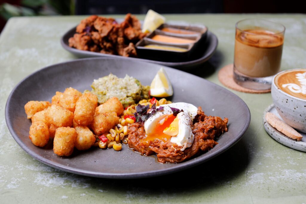 The first image shows a barista preparing drinks behind a glowing, tile-fronted coffee bar equipped with a professional espresso machine and warm pendant lights. The second image features a vibrant brunch spread with tater tots, a poached egg over pulled meat, and several coffee drinks served on a textured green table.