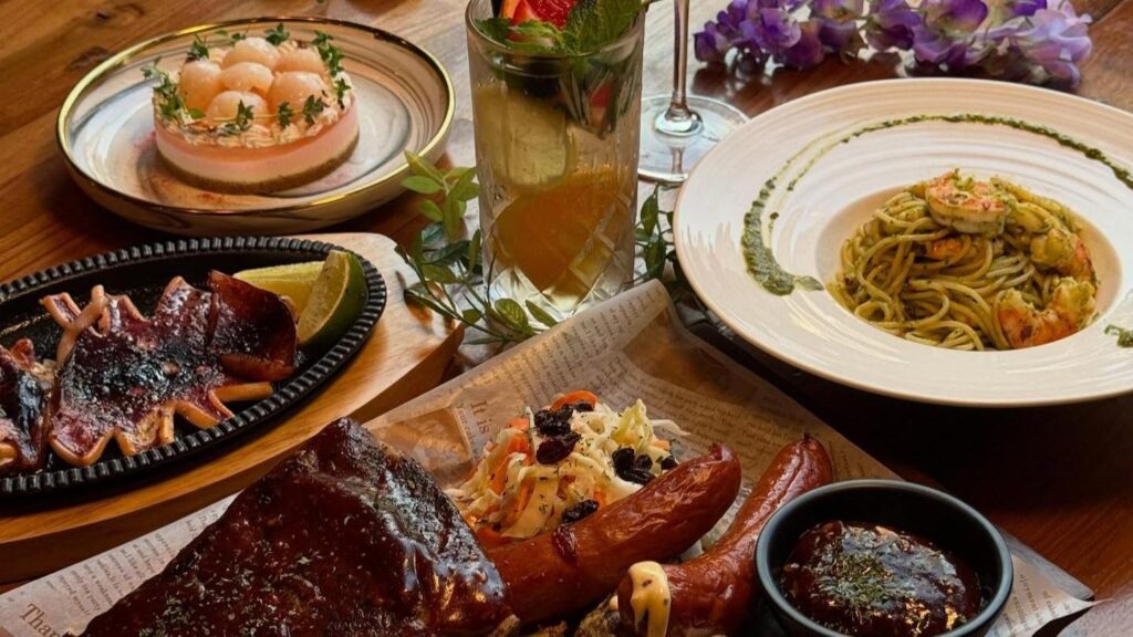 A vibrant spread of dishes including BBQ ribs, sausages, shrimp pesto pasta, and a lychee-topped dessert is arranged on a wooden table. A refreshing fruit-infused cocktail and purple flowers complete the aesthetic dining scene.