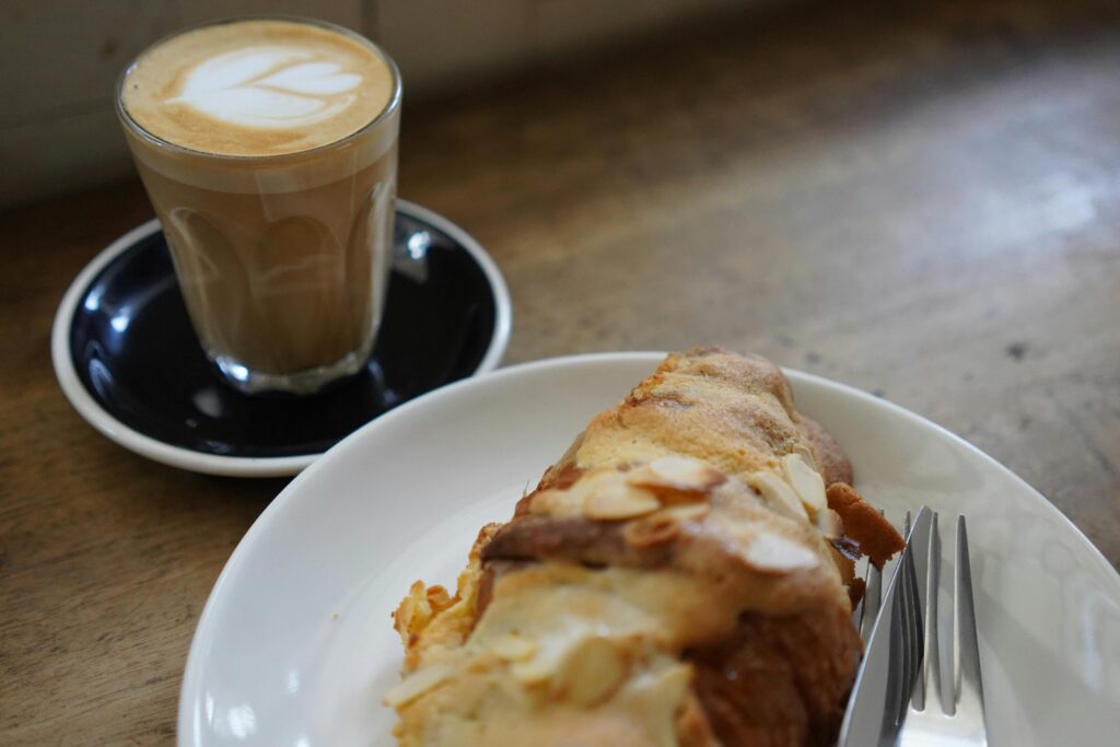 A glass of latte with heart-shaped latte art sits on a black saucer next to a white plate. The plate holds a golden-brown almond croissant topped with sliced nuts and a silver fork