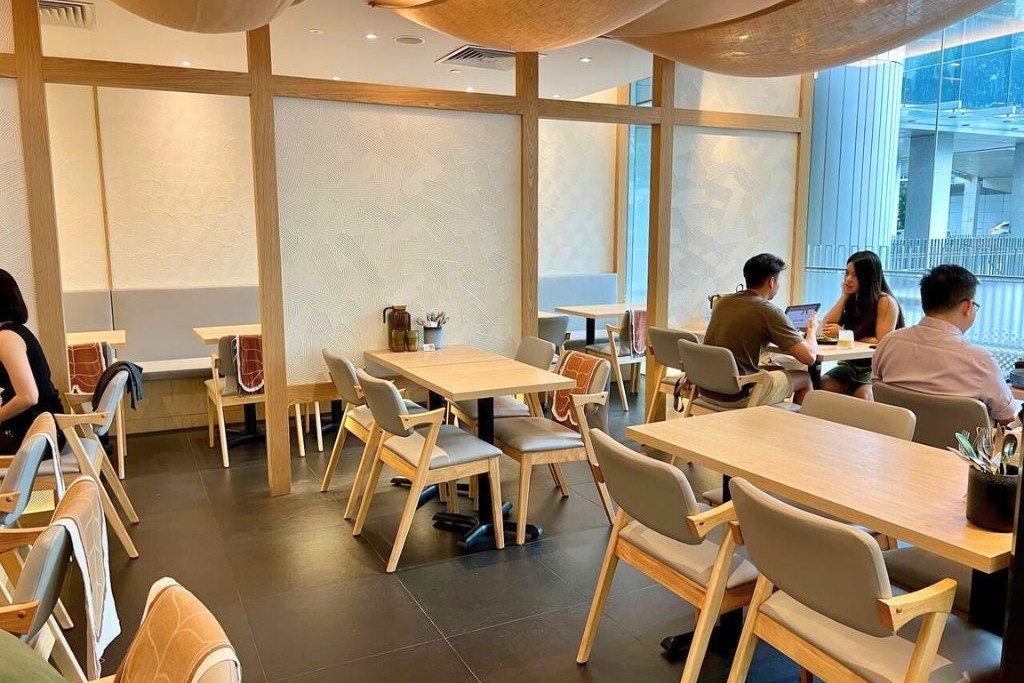 The image shows the interior of a modern, minimalist restaurant featuring light wood furniture, textured white walls, and warm ambient lighting. Several patrons are seated at tables, including a couple engaged in conversation and others working or dining in the clean, airy space.