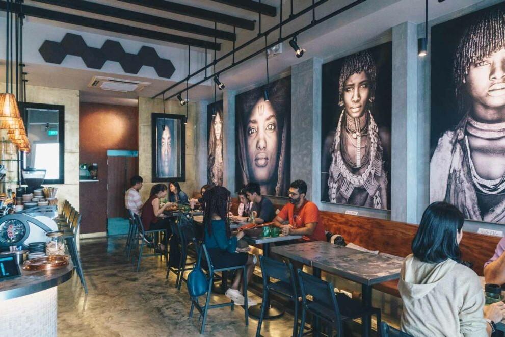 The dining area features large-scale black and white portraits of people on the walls, overlooking customers seated at tables and along a wooden banquet bench. The industrial-style space is characterized by exposed ceiling beams, track lighting, and a view of the coffee bar on the left.