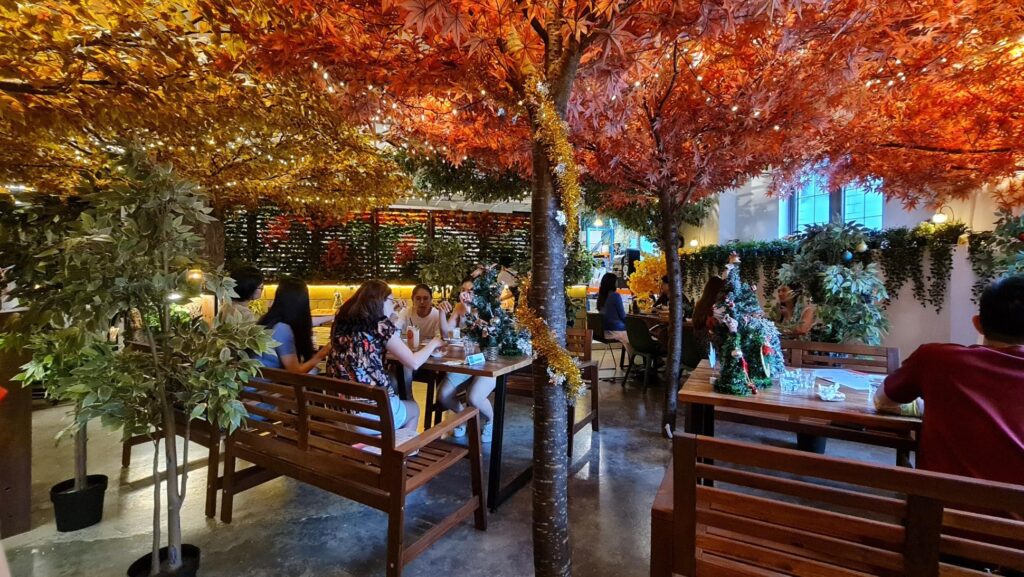 Patrons dine at wooden tables beneath a canopy of vibrant, artificial autumn leaves and festive fairy lights. The warm, indoor setting is further decorated with small Christmas trees and lush greenery to create a cozy, seasonal atmosphere.