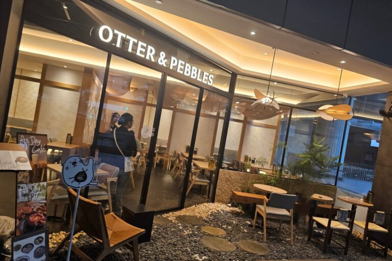 This image captures the exterior of a cafe named "OTTER & PEBBLES," featuring a sleek glass storefront and a distinctive pebble-covered outdoor seating area. Inside, the warm lighting and minimalist wood decor create a cozy, inviting atmosphere for patrons.