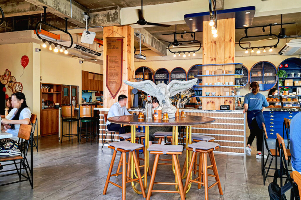 Industrial-style Singapore coffee shop interior with communal wooden tables, exposed concrete ceiling, and cozy specialty cafe atmosphere with customers seated indoors