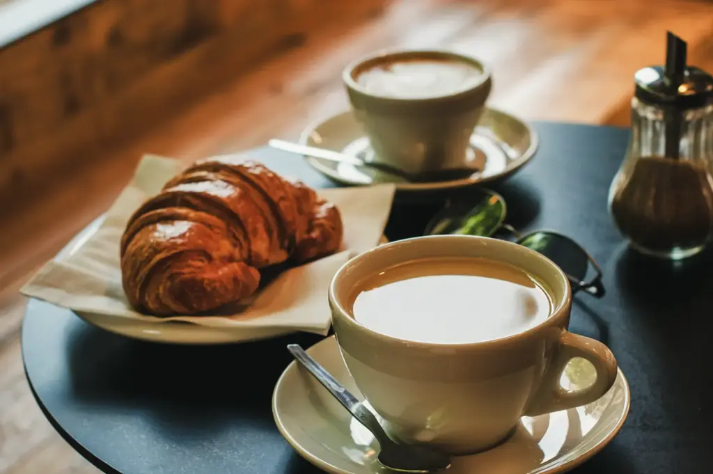 A croissant and two cups of coffee are on a black table, with a sugar dispenser and sunglasses nearby.