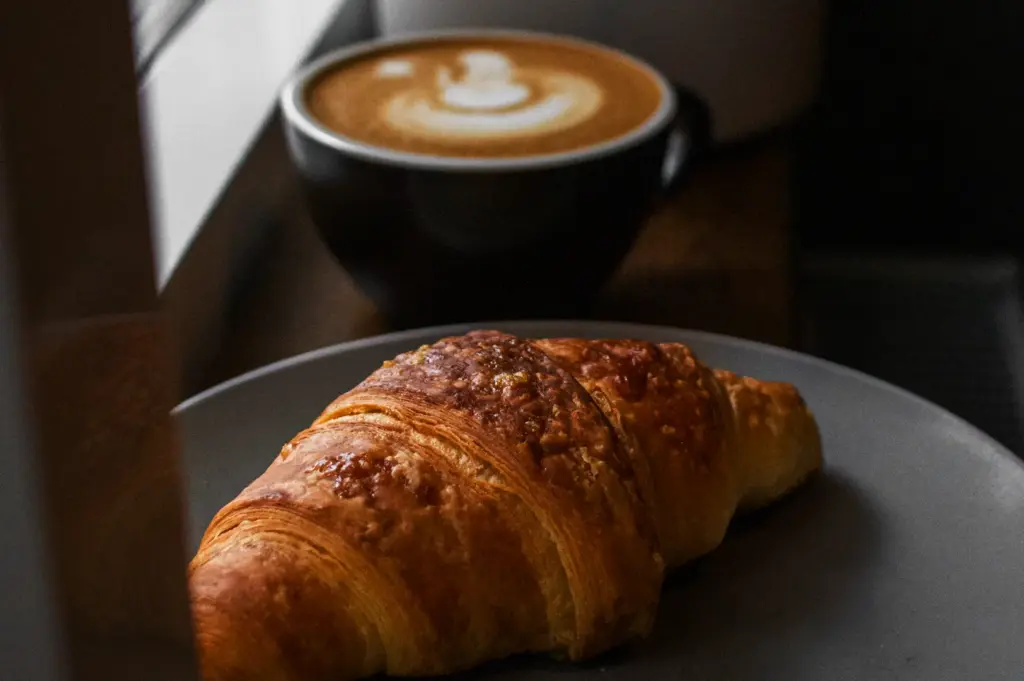 A golden-brown croissant is on a grey plate, with a cup of latte art coffee in the background.