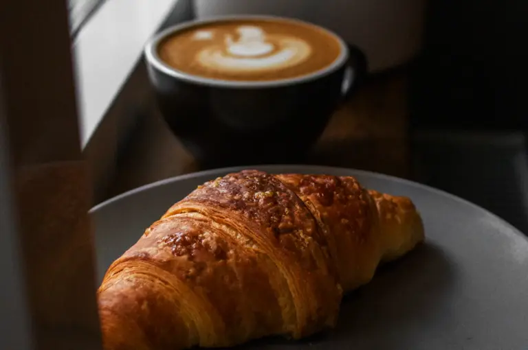 A golden-brown croissant is on a grey plate, with a cup of latte art coffee in the background.