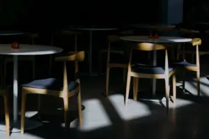 A dimly lit restaurant interior with round tables and wooden chairs, some with dark cushions. Sunlight casts dramatic shadows across the floor.