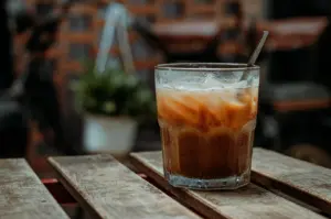 A glass of iced coffee with a straw sits on a wooden table. In the background, a potted plant and a brick wall are visible.